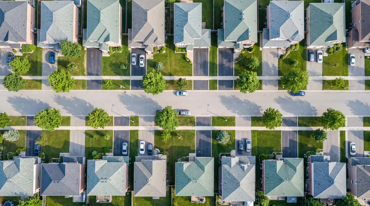 An aerial view looking straight down at a suburban residential street, rows of houses with driveways, lawns and mature trees.